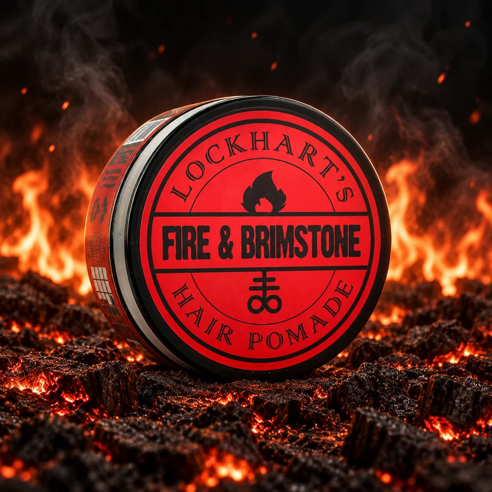 A round tin of Lockhart's Fire & Brimstone Hair Pomade sits on glowing embers with flames in the background. The tin has a bold red and black label with the brand name and product name clearly visible.